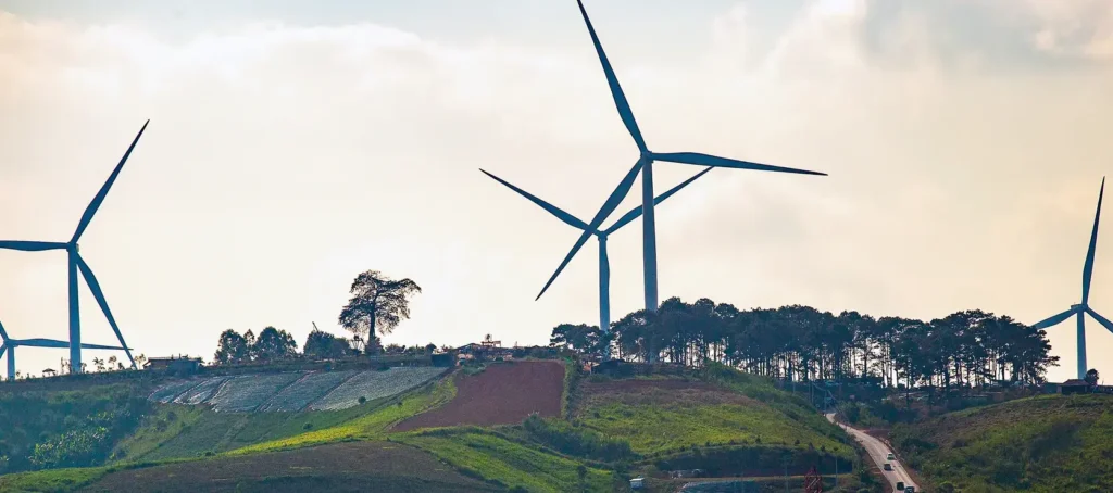Wind turbines on a green hillside surrounded by trees and farmland under a cloudy sky.