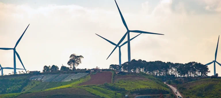 Wind turbines on a green hillside surrounded by trees and farmland under a cloudy sky.