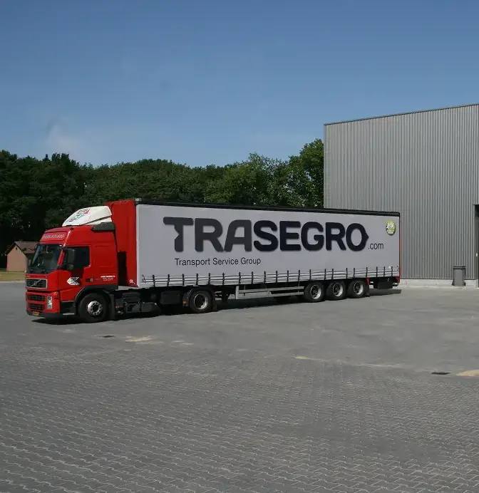 Red truck with a TRASEGRO.com trailer parked on a gray paved lot near a warehouse.